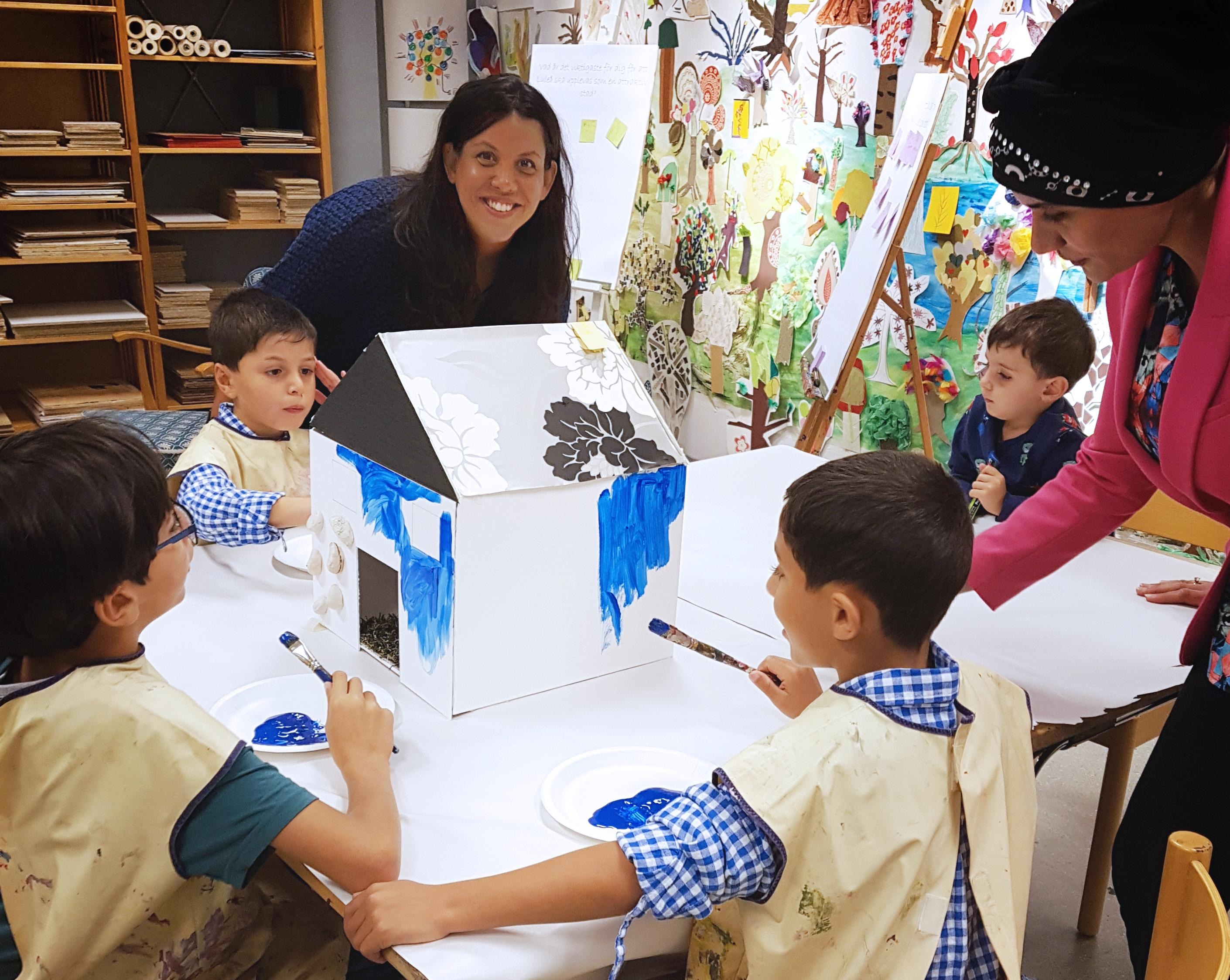 Barn och vuxna runt ett bord. Barnen målar på ett miniatyrhus.