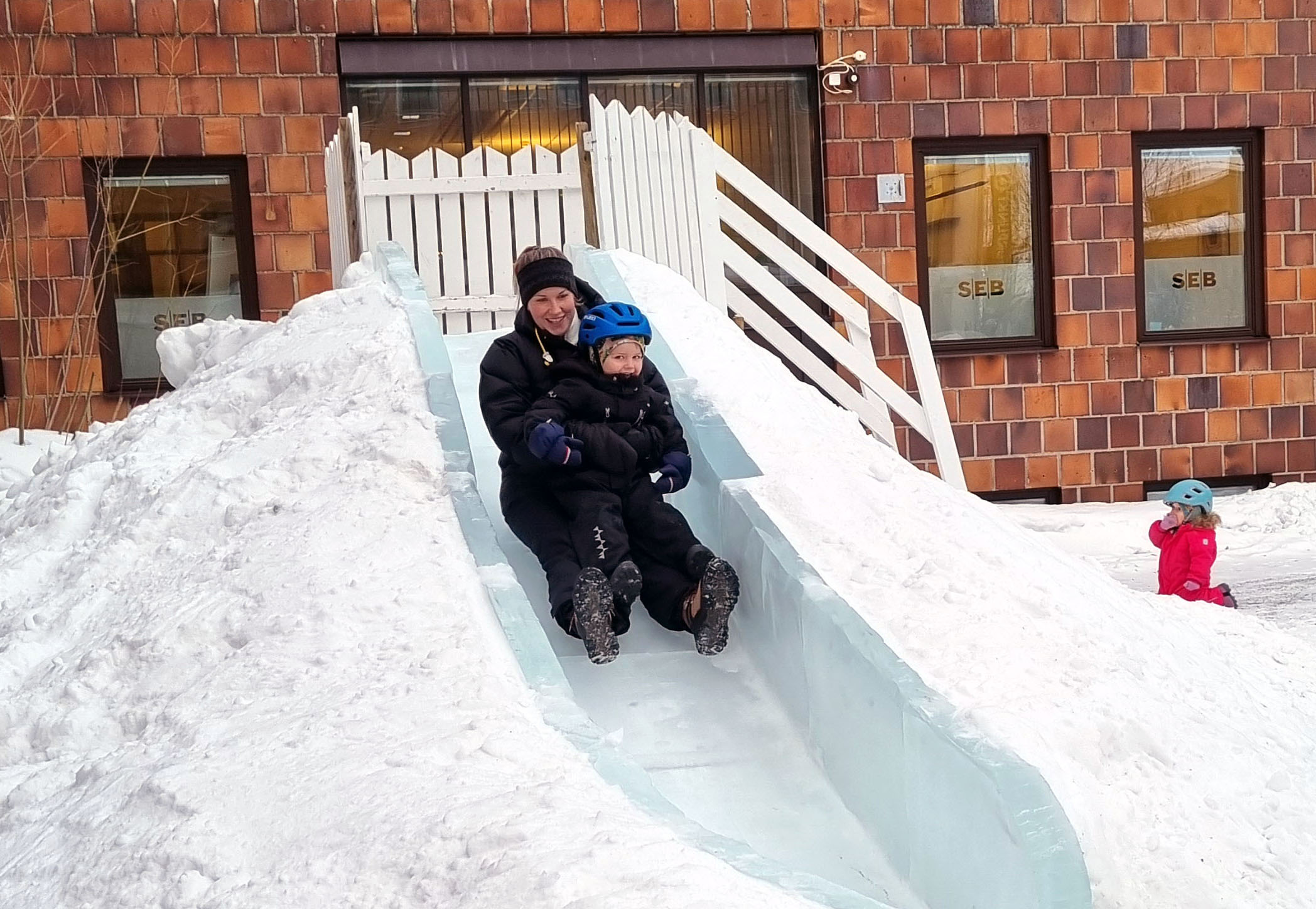 Tryggt sitter en liten pojke i mammas knä på väg nedför isrutschkanan. Lillasyster väntar på sin tur nedanför trappen upp till kanans topp.