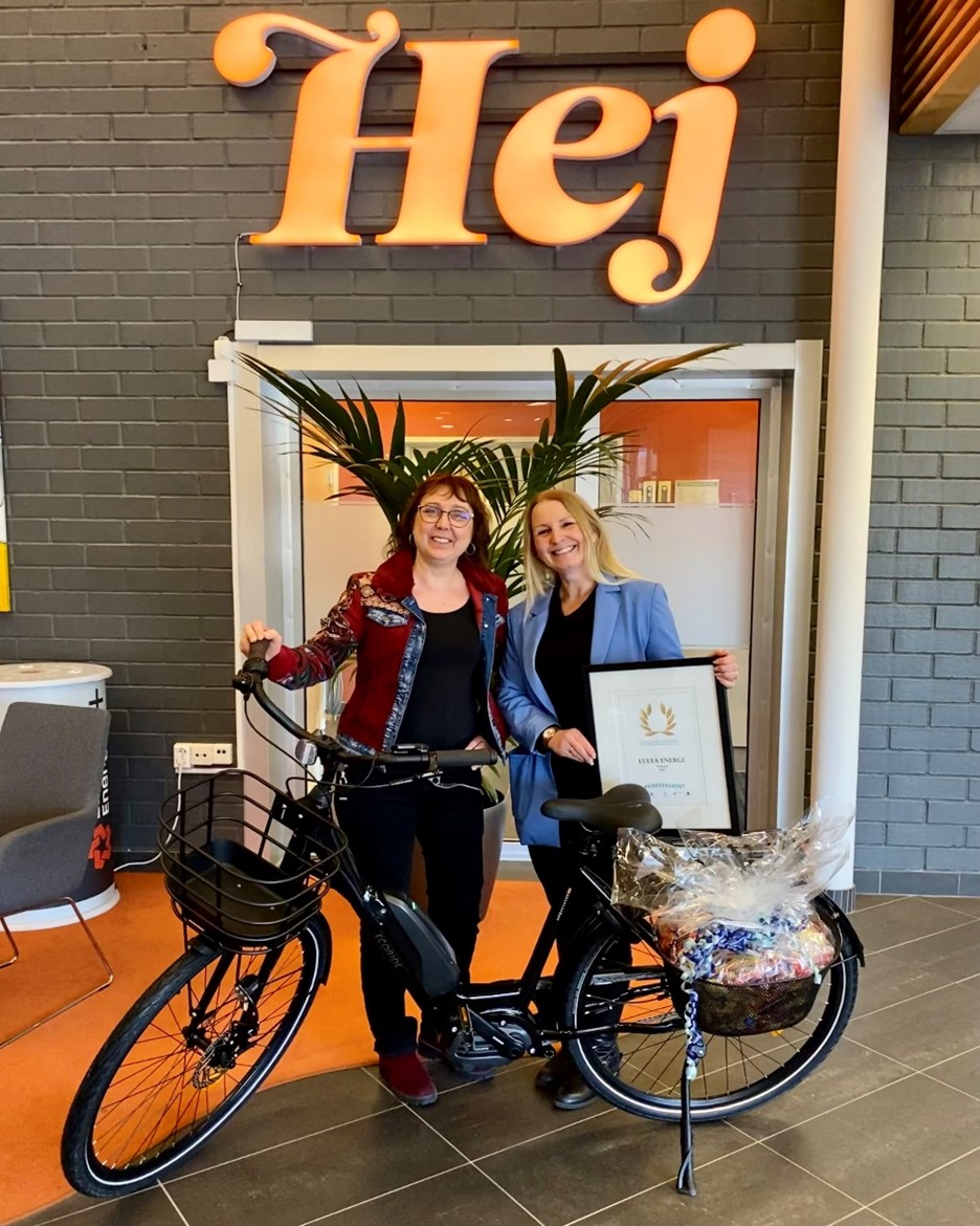 Glada vinnare – Katarina Kylefors och Sandra Östling, Luleå Energi. De står bakom vinsten som är en elcykel. På väggen bakom står ett stort Hej.