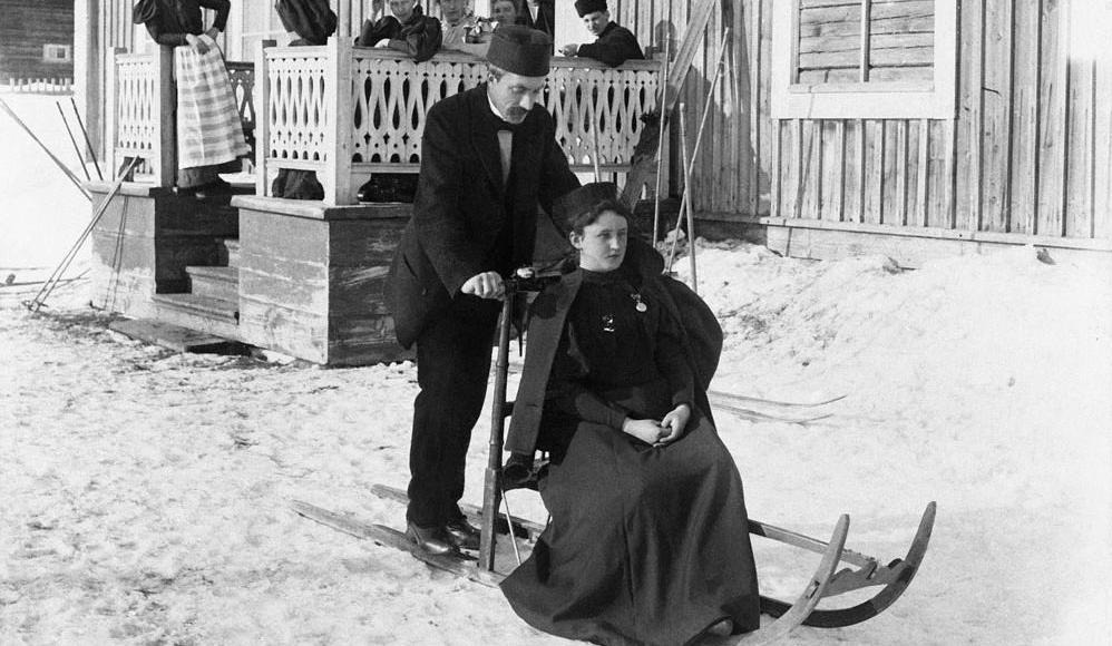 Bilden visar en kvinna i lång kjol som sitter på en spark. Bakom henne står en man i kostym med cylinderformad mössa. I bakgrunden skymtar en farstukvist med folk på.