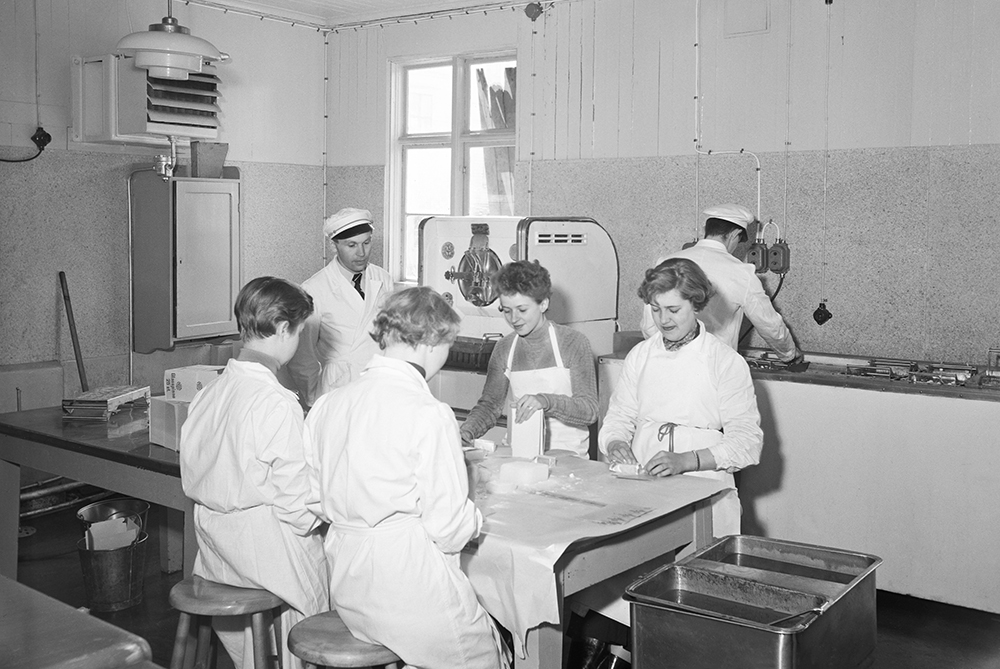 Vitklädda kvinnor sitter och slår in glasspinnar för hand. Två män i keps som tittar på respektive man som arbetar vid en bänk.
