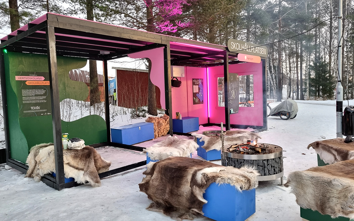 Skolhållplatsen ser ut som en stor busshållplats med stålram och glasrutor. Delar av glasväggarna är heltäckta med rosa skivor eller gröna skivor. Lampor lyser upp innuti och det finns bänkar med renskinn på. Utanför finns fler bänkar med renskinn och en rund grillplats. På en vägg finns en griffeltavla där man kan skriva med kritor.