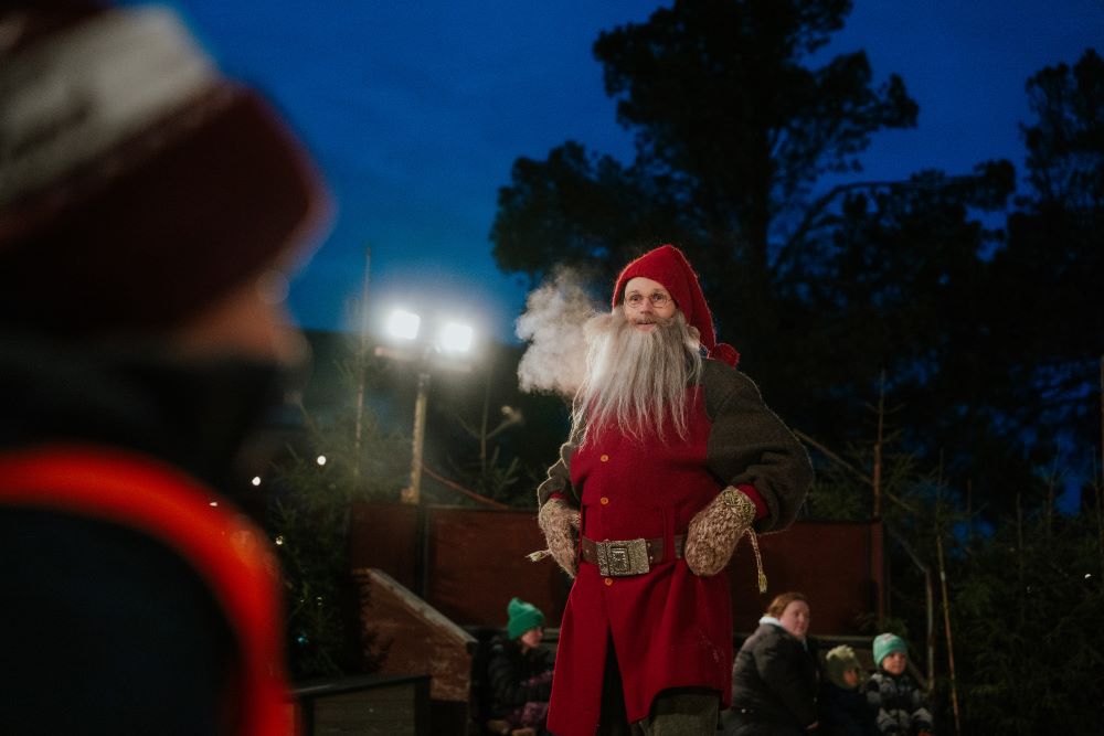 Tobias Carlsson spelar Tomten i julkalendern i Stadsparken. Han är klädd i röda kläder och tomteluva. Han står med händerna i sidorna. Publiken syns i förgrunden och i bakgrunden.