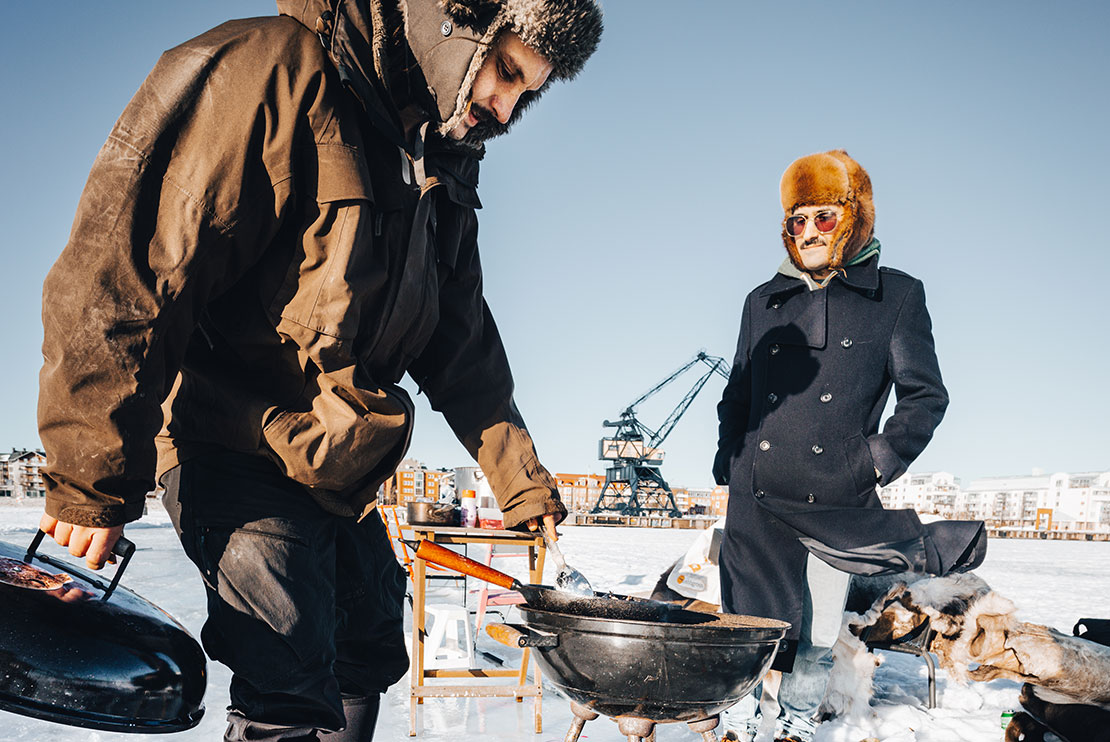 Två vinterklädda män grillar mat över öppen eld ute på isen.