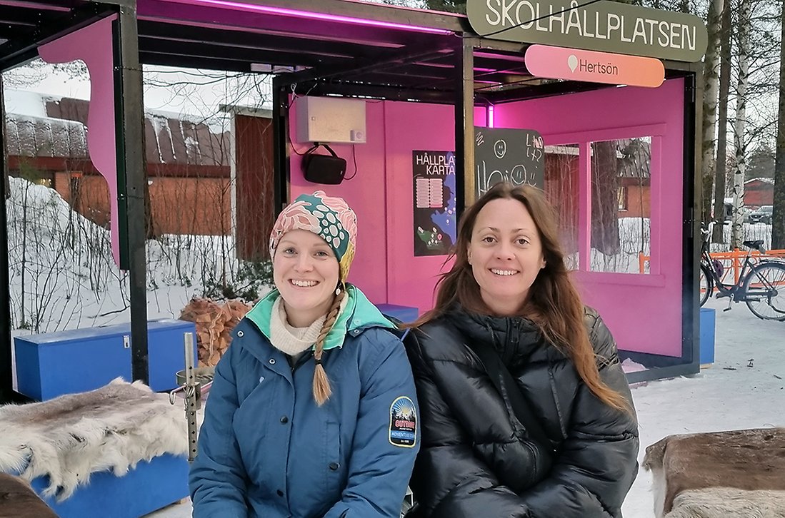 Jessica Rhodin i ljusblå jacka och mönstrad mössa och Ida Lemoine i svart jacka och utsläppt mörk hår sitter på renskinn framför skolhållplatsen. I bakgrunden brinner en eld.