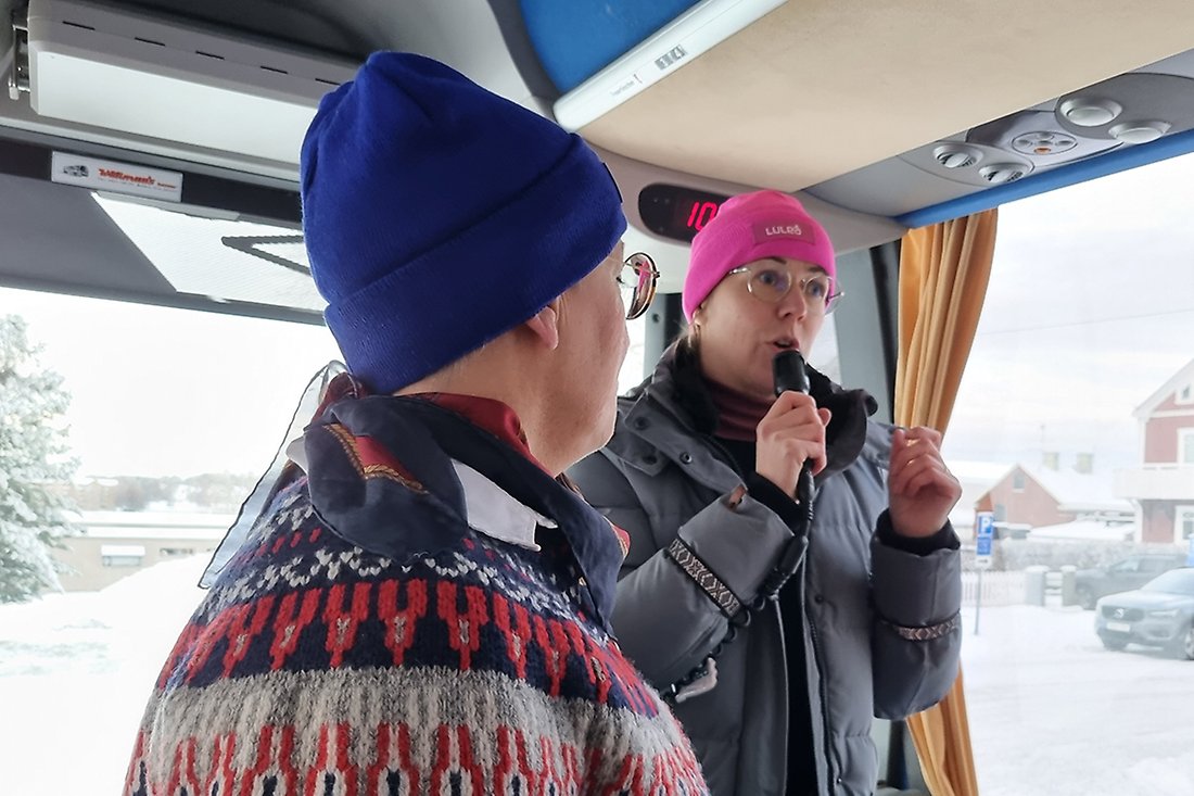 Två kvinnor med mössor står längst fram i en buss. Den ena pratar i mikrofon.